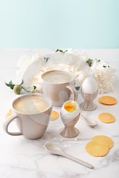 Soft-boiled eggs with liquide orange yolk in ceramic egg cups, two cups of coffee with ceramic spoons and thin crispy