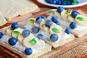 Soft cheese cream, bananas and berries sandwiches. Cracker bread with toppings on wooden board. Closeup