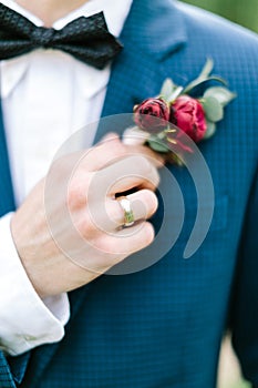 Soft focus of cropped groom`s hand in wedding blue tuxedo and bowtie gently touches a boutonniere made of red peonies