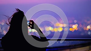 Soft focus and Silhouette of young woman with long hair to take a photo cityscape on top of building with city colourful bokeh lig