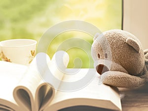 Soft focus of Teddy bear laying on open book like heart shape page, A shape of heart on book with teddy and cup of tea next to win