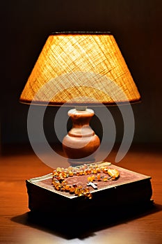 Soft light of table lamp, old book prayer book or Bible and amber Catholic rosary on table. Religion, evening prayer. Religion