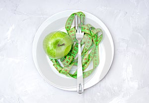 Soft measuring tape, fresh green apple and a fork on a white plate on a concrete table, top view.