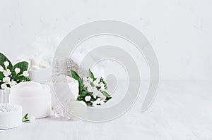 Soft pure white delicate cosmetics set of cream, salt, clay decorated white flowers, green leaves on light soft wooden background.