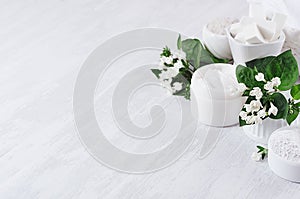 Soft pure white delicate cosmetics set of cream, salt, clay decorated white flowers, green leaves on light soft wooden background.