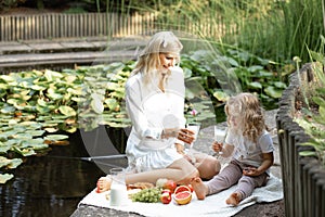 Soft, smiling blond woman and little girl drink milk from glass on picnic, organic fresh fruit and dairy lunch near lake