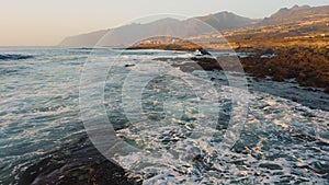 Soft sunset light reflected in the ocean water and illuminates rocky shore. Dark sea in evening. Foaming waves crashing