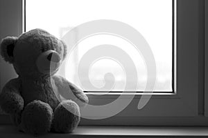 A soft toy a sad bear sits on a windowsill. There are noises in the photo, because the photo is dark