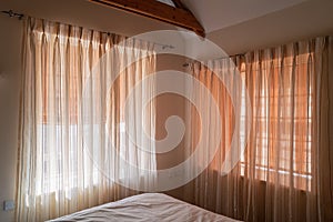 Soft translucent curtains hanging on two windows in a bedroom that has wood beams. There are blinds behind the net type curtains