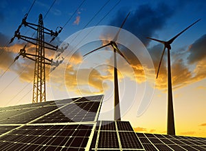 Solar panels with wind turbines and electricity pylon at sunset.