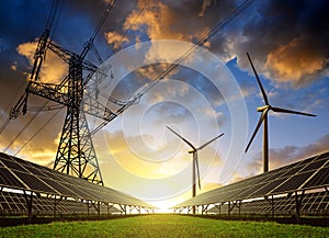 Solar panels with wind turbines and electricity pylon at sunset.