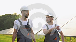 Solar power plant with two engineers in protective workwear with tools walking and examining photovoltaic panels