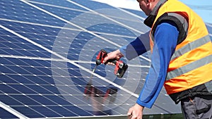 Solar power plant worker standing on a stepladder tightens solar panels fixing bolts with a drill.