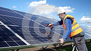 Solar power plant worker standing on a stepladder tightens solar panels fixing bolts with a drill.