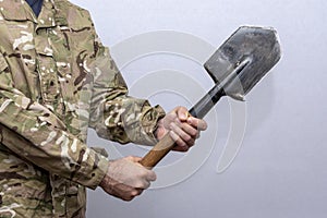 A soldier in an American military uniform holds a sapper shovel in his hands on a light background.