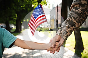 Soldier and his little son with American flag holding hands outdoors, closeup. Veterans Day in USA