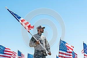 Soldier in military uniform and cap holding american flag while standing against blue sky