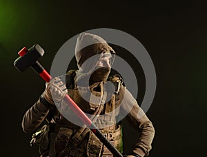 A soldier-saboteur rebel in military clothing with a weapon on a dark background holding a sledgehammer