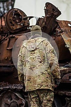 Soldier in uniform near a burnt and melted rusty wreckage of a Soviet Russian-made tank