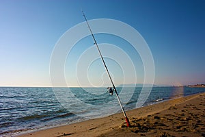 A solitary fishing rod rises from the ground on which it is planted on the sand of the beach and stands against the blue ocean sea