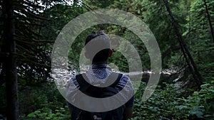 A solitary middle aged man with a backpack looking out at a river in an evergreen forest.