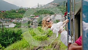 Solo female traveler leans out train door, wind in curly hair. Smiling woman enjoys tropical scenery on rail journey