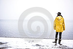 Solo traveler with yellow rain coat and cowbow hat lookng away under snow, back shot