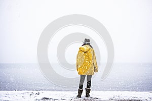 Solo traveler with yellow rain coat and cowbow hat lookng away under snow, back shot