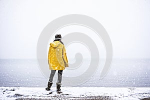 Solo traveler with yellow rain coat and cowbow hat lookng away under snow, back shot