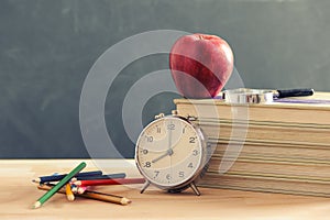 Some books and a pencil holder on a wooden table. Red apple is standing on the books.