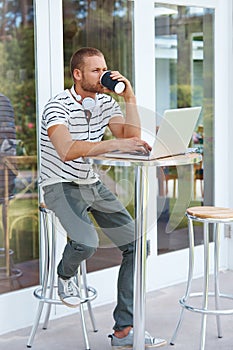 Some caffeine to fuel his imagination. a handsome young man working on a laptop outside and drinking some coffee.