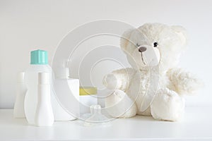 Some cosmetic, perfume and lotion bottles on a white table next to a teddy bear and a pacifier.