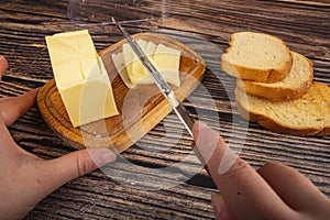 Someone cuts some butter with a knife from a piece of butter in a wooden butter dish and fresh wheat toast on a wooden background