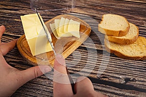 Someone cuts some butter with a knife from a piece of butter in a wooden butter dish, slices of cheese and fresh wheat toast on a