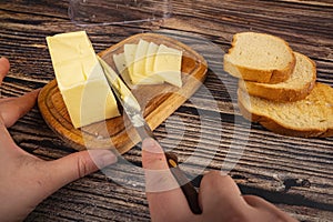 Someone cuts some butter with a knife from a piece of butter in a wooden butter dish, slices of cheese and fresh wheat toast on a