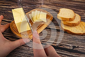 Someone cuts some butter with a knife from a piece of butter in a wooden butter dish, slices of cheese and fresh wheat toast on a