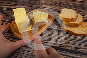 Someone cuts some butter with a knife from a piece in a wooden butter dish and fresh wheat toast on a wooden background. Close up