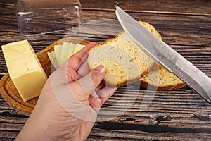 Someone spreads butter on fresh wheat toast with a knife, a wooden butter dish with a piece of butter and slices of cheese on a