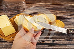 Someone spreads butter on fresh wheat toast with a knife and a wooden butter dish with a piece of butter and slices of cheese on a