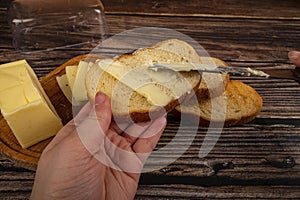 Someone spreads butter on fresh wheat toast with a knife and a wooden butter dish with a piece of butter and slices of cheese on a