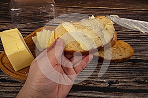 Someone spreads butter on fresh wheat toast with a knife and a wooden butter dish with a piece of butter and slices of cheese on a
