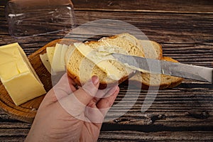 Someone spreads butter with a knife on fresh wheat toast, a piece of butter and slices of cheese in a wooden butter dish on a