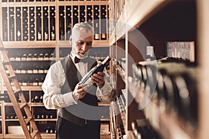 Sommelier Concept. Senior man standing taking wine from cabinet reading curious