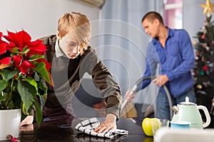 Son cleans the table with a rag while father vacuums room before christmas