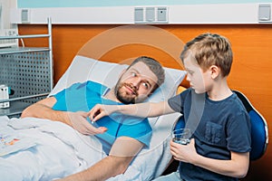 Son giving pills and glass of water to his sick father laying at ward