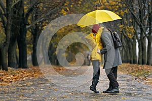 Son are hugging his mom walking in the autumn park in rain with large yellow umbrella. Rainy day