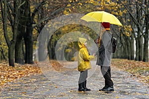 Son with mom walking in the autumn park in rain with large yellow umbrella. Rainy family day