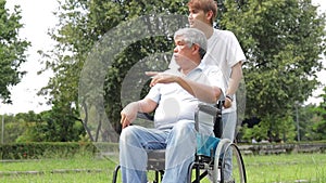 A son takes his father, who is in a wheelchair, for a walk in an outdoor park.