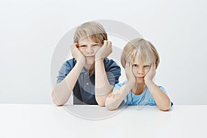 Sons grounded for bad behaviour at school. Bored unhappy cute male children sitting at table, leaning heads on hands and