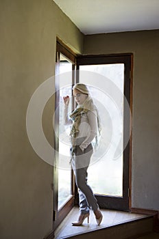 Sophisticated blonde woman wearing white shirt, grey pants,heels and scarf, posing next to glass doors with the light streaming in